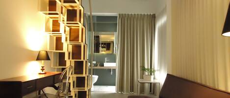 A modern hotel room with a staircase, a desk, a sofa, a small table with a plant, and a bathroom visible through a door.