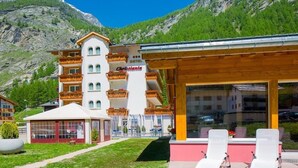 A hotel with a white facade, multiple balconies, and a wooden structure with lounge chairs. - Christiania Wellness Hotel (Saas Almagell)
