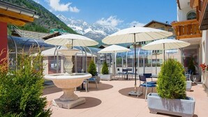 An outdoor patio with tables and chairs, umbrellas, and a fountain. - Christiania Wellness Hotel (Saas Almagell)
