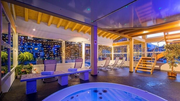 A modern indoor pool area with a hot tub, seating, and a view of the cityscape through large windows. - Christiania Wellness Hotel (Saas Almagell)