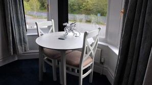 Een eethoek met een witte tafel en vier stoelen, een decoratieve draagbeeldjes, en uitzicht op een weg en groen buiten.