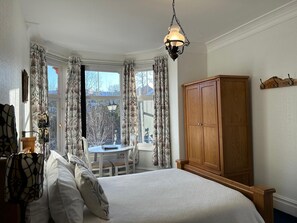 A bedroom with a bed, a wooden wardrobe, a table with chairs, and a view through a window with curtains.