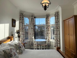 A bedroom with a bed, a wooden wardrobe, a table with chairs, and a view through large windows.
