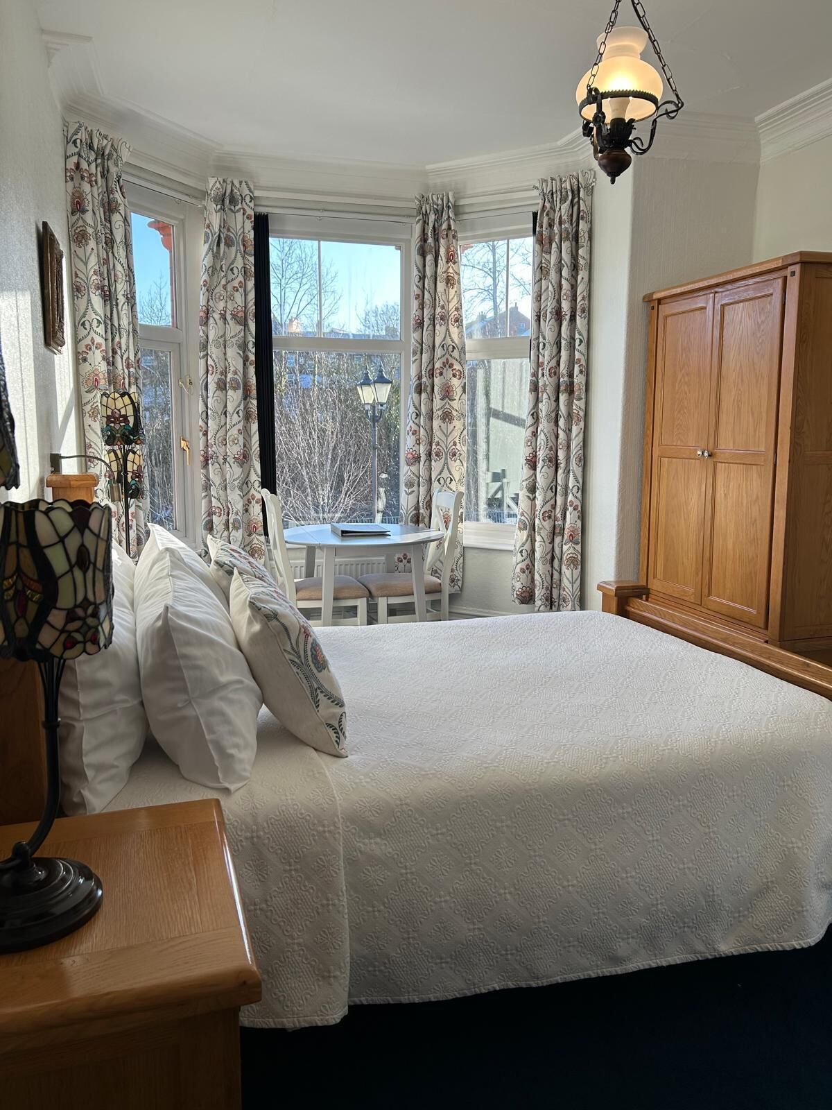 Une chambre à coucher avec un grand lit, une armoire en bois, un bureau avec une lampe et une fenêtre avec des rideaux.