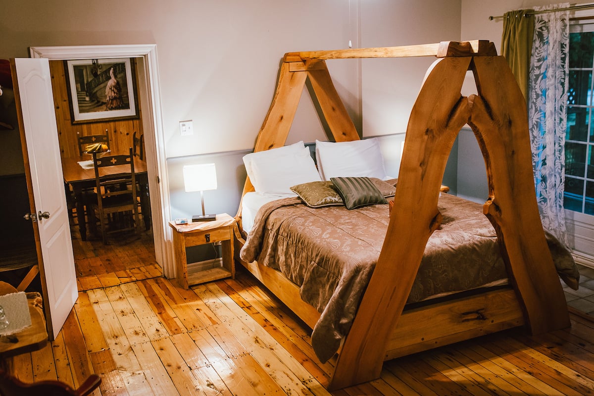 A rustic wooden bed with a canopy, a bedside table with a lamp, and a chair in a room with wooden flooring and a door leading to another room.