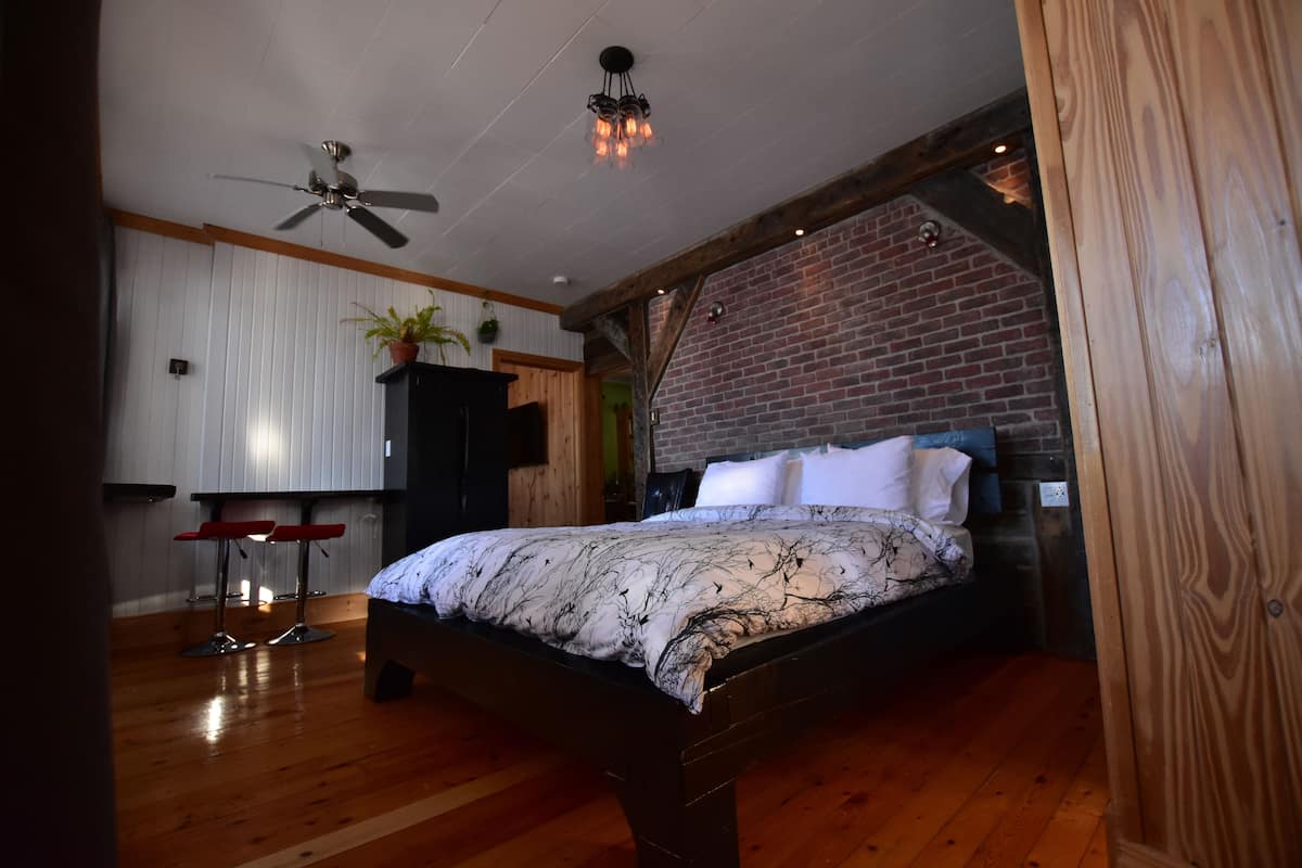 A bedroom with a wooden bed, a ceiling fan, a brick wall, and a small kitchenette area.
