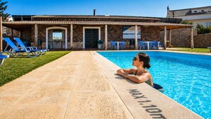 A swimming pool with a person relaxing by the edge, a covered patio area with chairs, and a stone building with a tiled roof. - Tonel Apartments (Vila do Bispo)