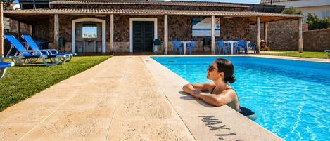 A swimming pool with a person relaxing by the edge, a covered patio area with chairs, and a stone building with a tiled roof.