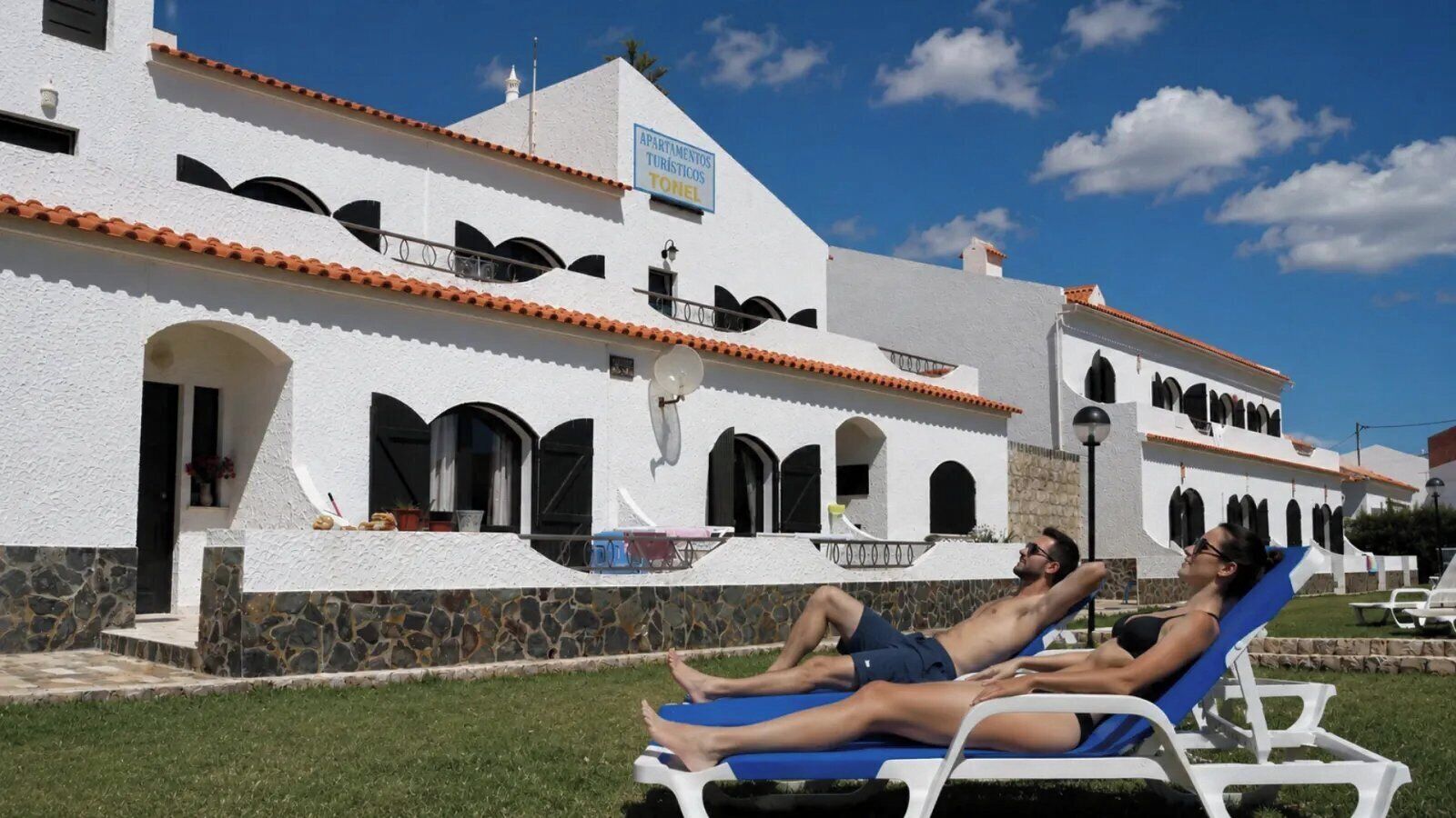 Un bâtiment blanc avec une enseigne indiquant « Appartements touristiques » et « Toilettes », signalant un complexe d’appartements touristiques doté d’une piscine.