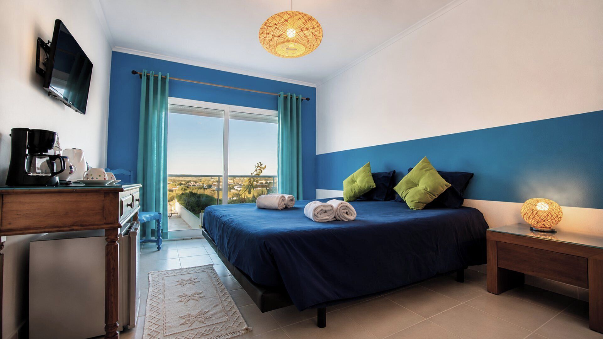 A bedroom with a bed, a desk with a coffee maker, a TV, and a balcony with curtains.