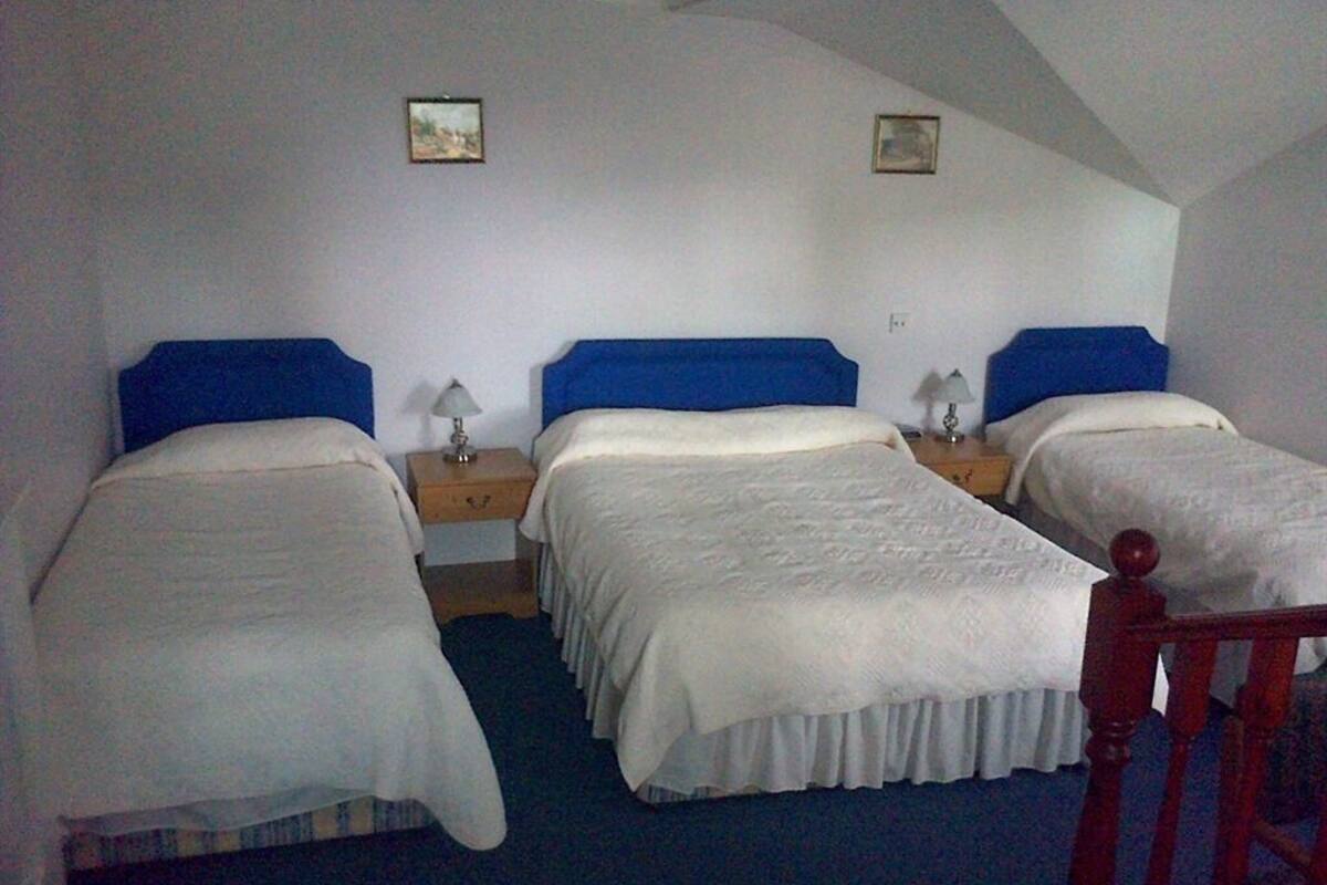 A room with three single beds, each with a blue headboard and white bedding. There are two bedside tables with lamps. The room has a sloped ceiling and two framed pictures on the walls.