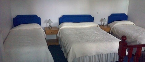 A room with three single beds, each with a blue headboard and white bedding. There are two bedside tables with lamps. The room has a sloped ceiling and two framed pictures on the walls.