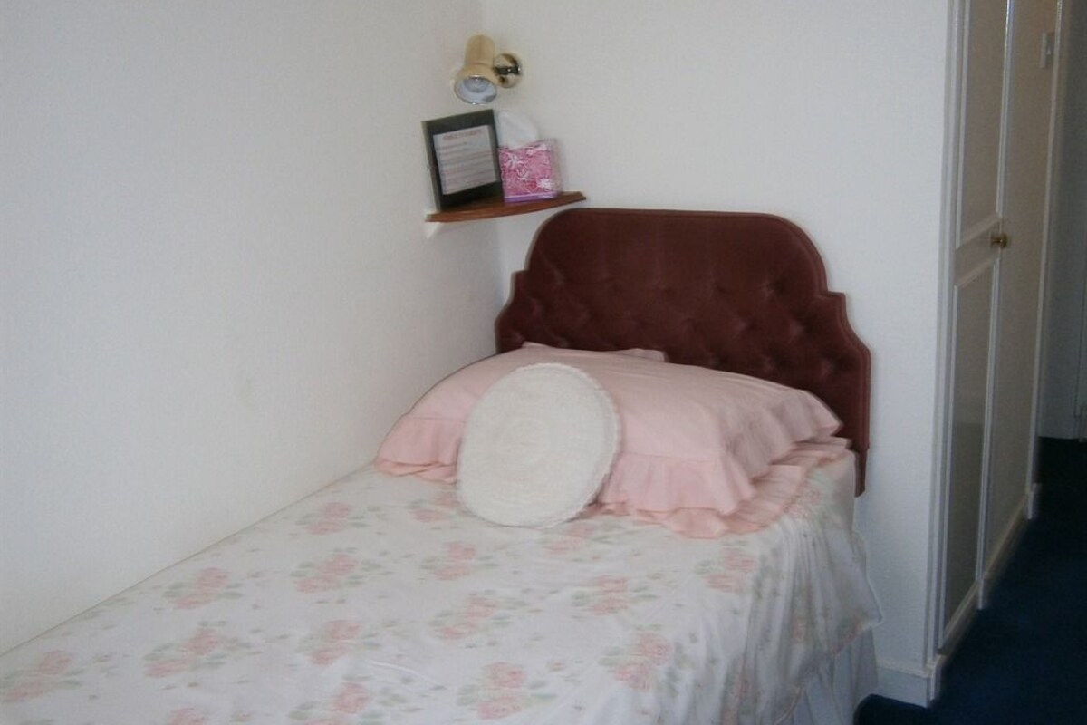 A small bedroom with a single bed, a nightstand, a wall-mounted shelf with a lamp, and a door.