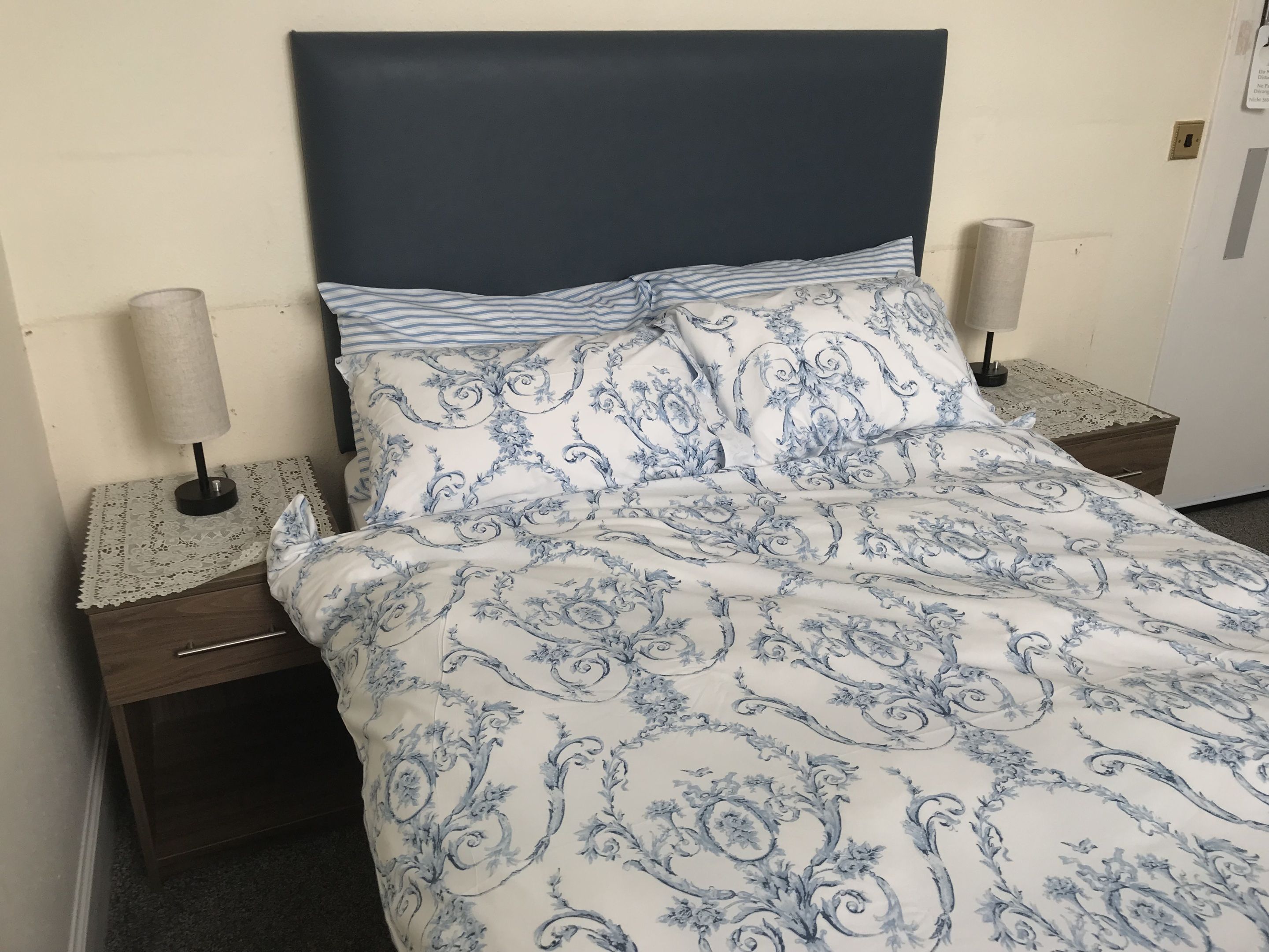 Un lit recouvert d’une couvre-lit à motifs floraux bleus et blancs, deux lampes de chevet et une table de chevet en bois avec un tiroir.