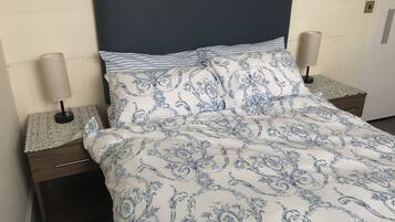 Un lit recouvert d’une couvre-lit à motifs floraux bleus et blancs, deux lampes de chevet et une table de chevet en bois avec un tiroir.
