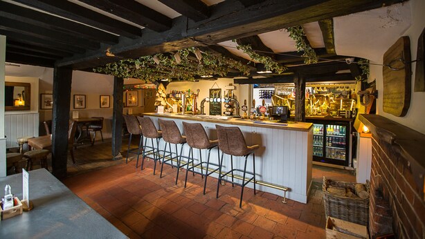 A bar area with a rustic wooden ceiling, bar stools, and a counter with a variety of bottles and glasses. - The Bull Inn Pub (Reading)