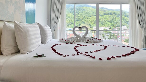 A hotel room with a bed, white curtains, and a view of greenery through large windows. - Patong Platinums (Patong)