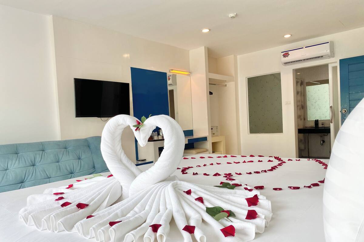 A hotel room with a bed adorned with a swan-shaped towel arrangement and rose petals, a blue sofa, a flat-screen TV, and an air conditioning unit.