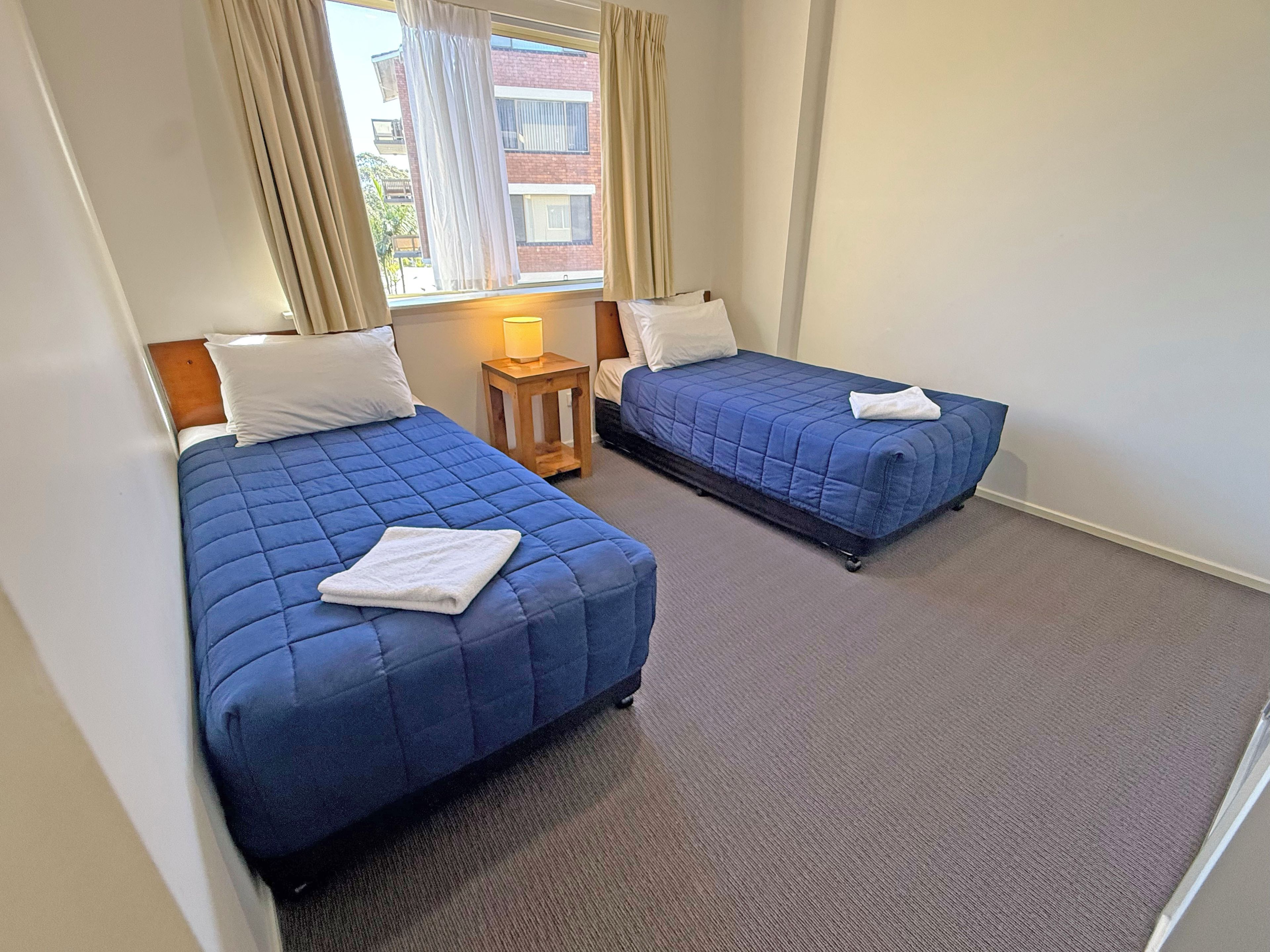 Two single beds with blue bedding, a small wooden nightstand with a lamp, and a view of a building through a window with curtains.