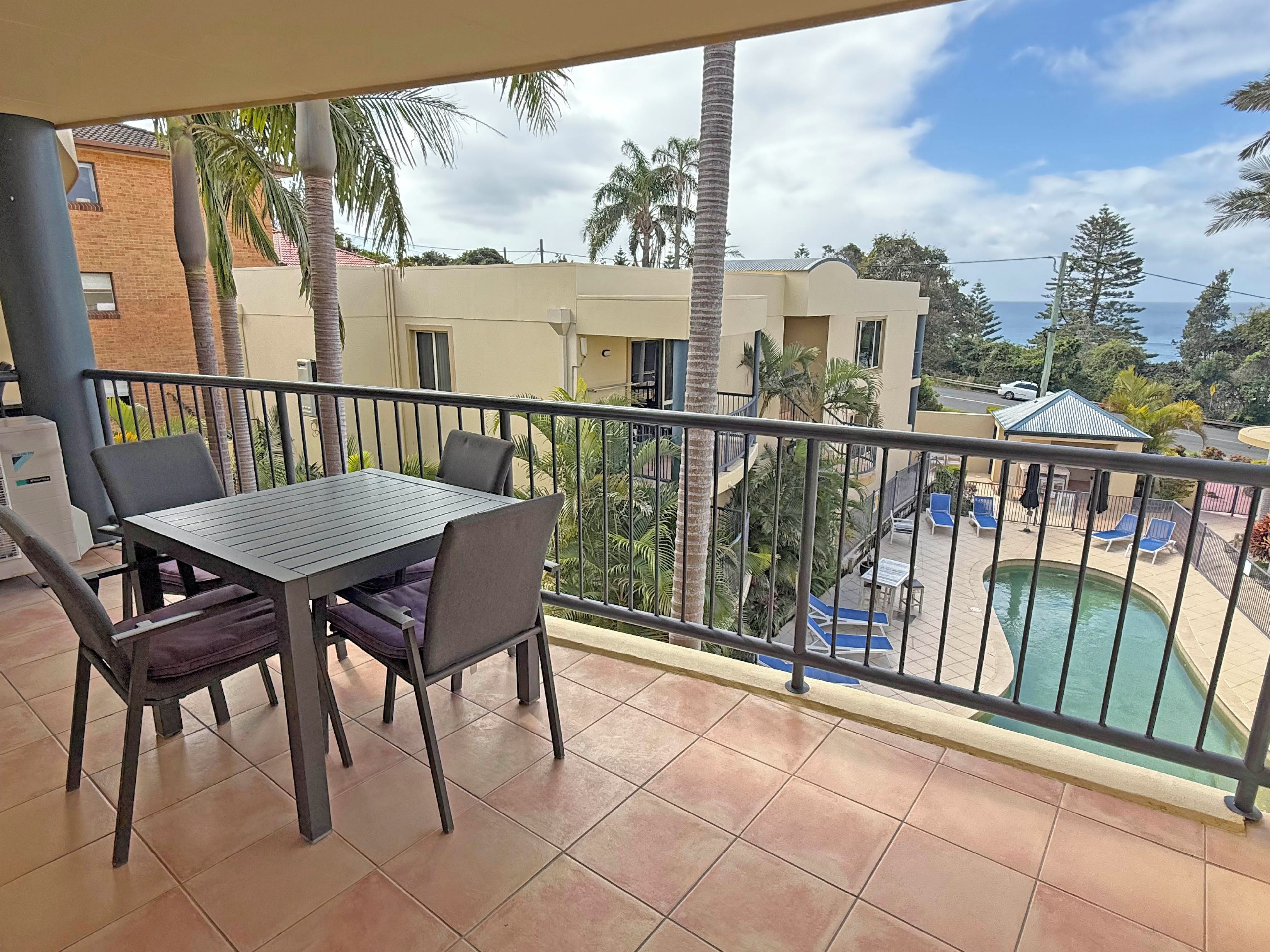 A balcony with a table and chairs, overlooking a swimming pool and a residential area with palm trees.