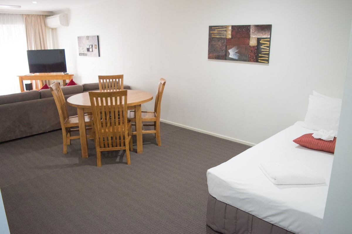 A hotel room with a bed, a dining table with chairs, a sofa, a TV, and artwork on the walls.