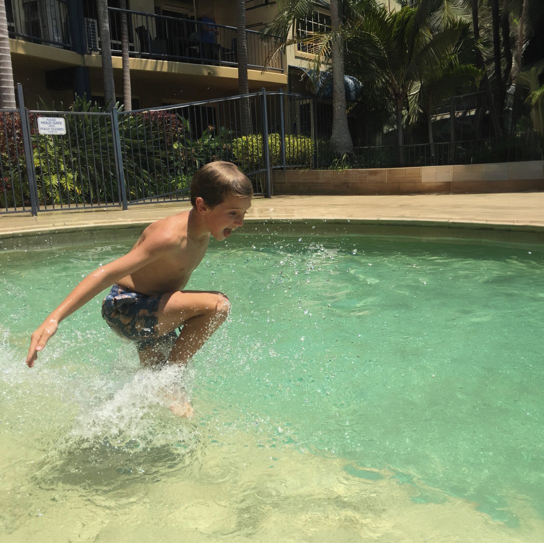 Zona de piscina con una persona en traje de baño, rodeada por una valla y palmeras.