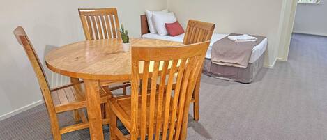A round wooden dining table with four chairs, a small potted plant, and a sofa with cushions in a room with carpeted flooring and a window.