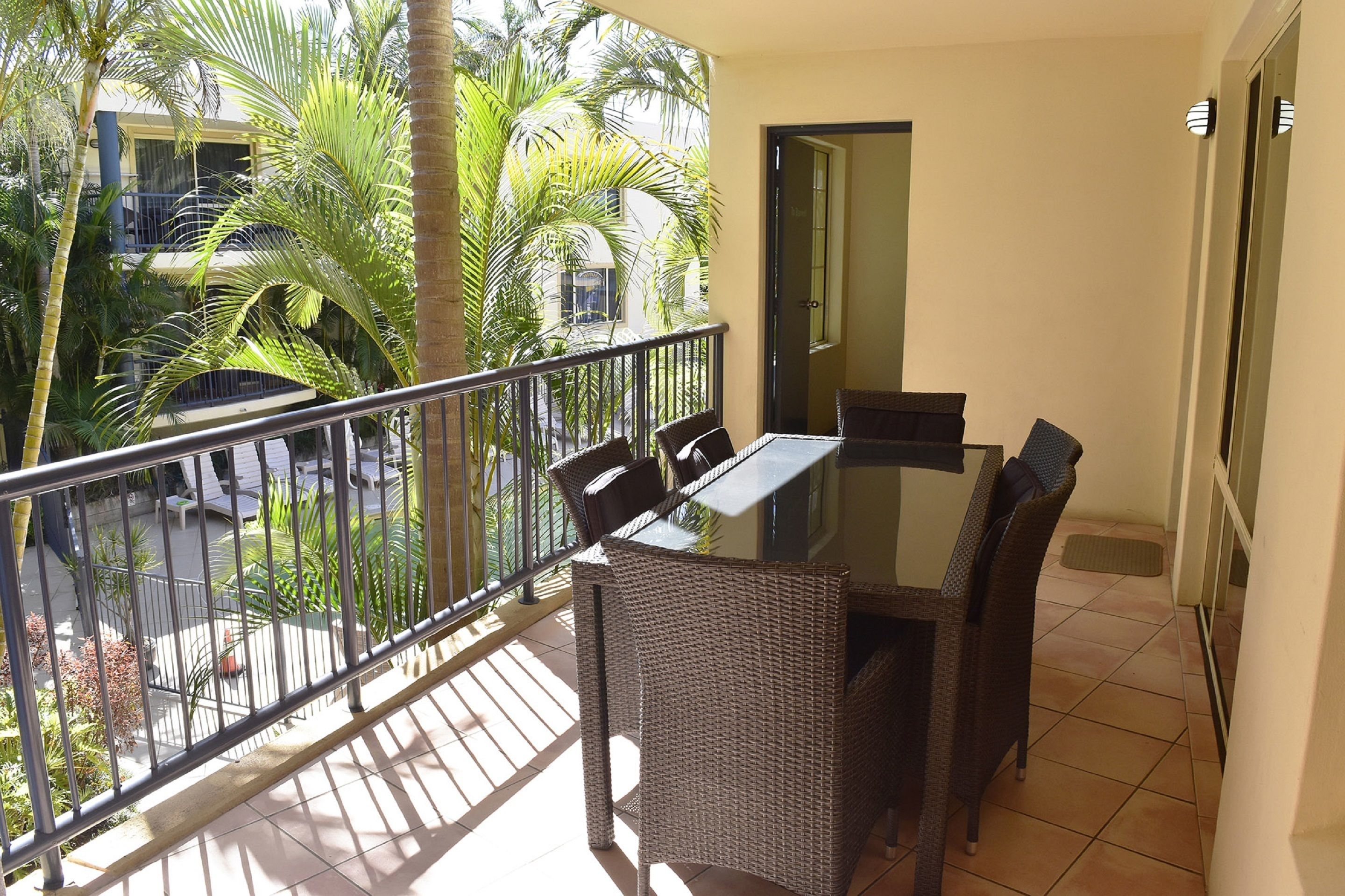 A balcony with a dining table and chairs, surrounded by a metal railing, overlooking a courtyard with palm trees and a building.
