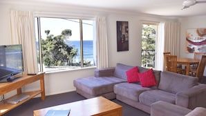 A living room with a grey sofa, a wooden coffee table, a TV, and a view of the ocean through large windows. - Beachside Holiday Apartments (Port Macquarie)