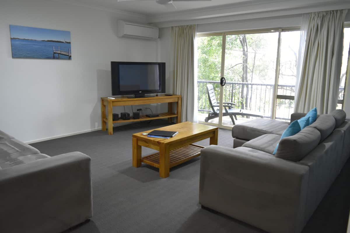A living room with a sofa, a coffee table, a TV on a wooden stand, and a balcony with a chair.