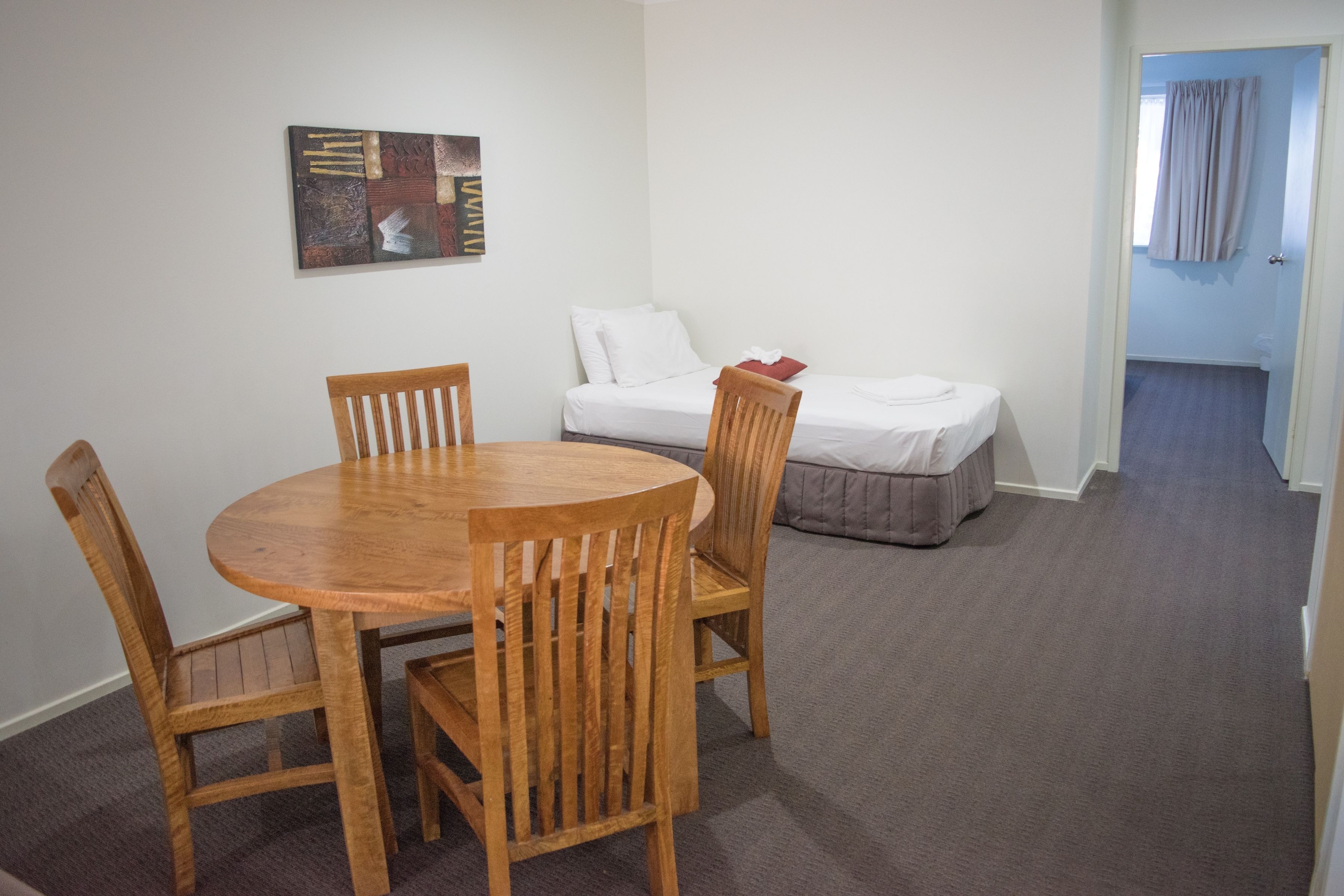 A room with a round wooden table and four chairs, a bed with white bedding, and a painting on the wall.