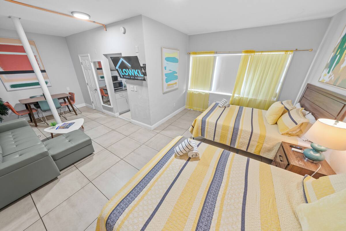 A modern hotel room with a bed, sofa, and a small table. There is a window with curtains, a door, and a sign on the wall.