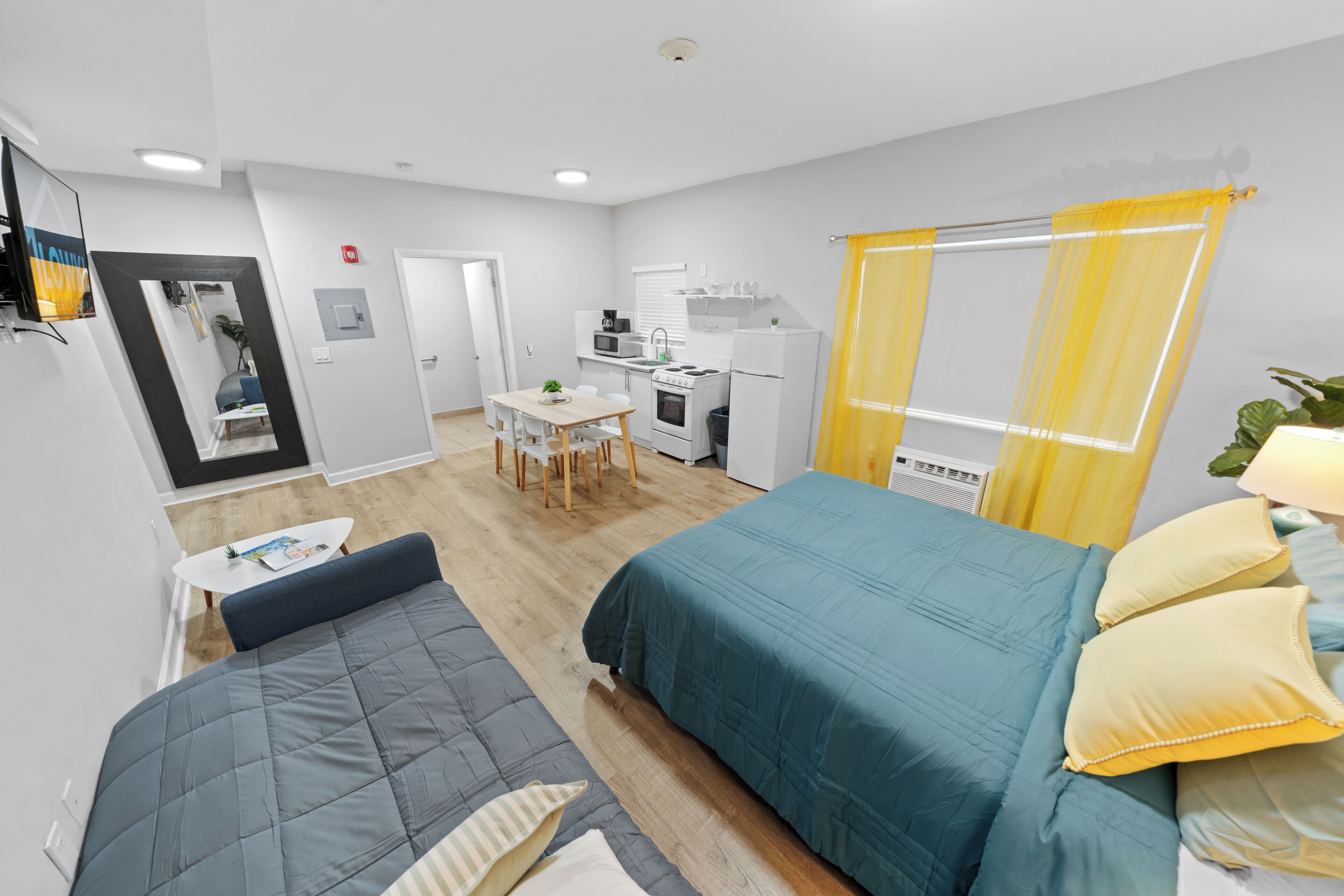 A compact hotel room with a bed, a small table, a sofa, a kitchenette, and a bathroom.