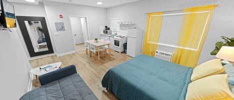 A compact hotel room with a bed, a small table, a sofa, a kitchenette, and a bathroom.