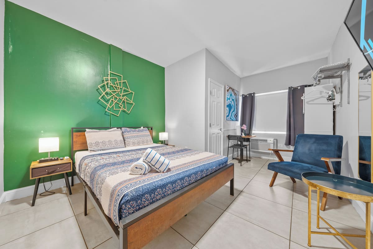 A bedroom with a green accent wall, a bed with patterned bedding, a blue chair, and a desk with a chair.
