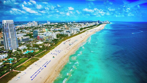 An aerial view of a coastal city with a long beach, high-rise buildings, and a clear blue sky. - Miami International by Lowkl (Miami Beach)