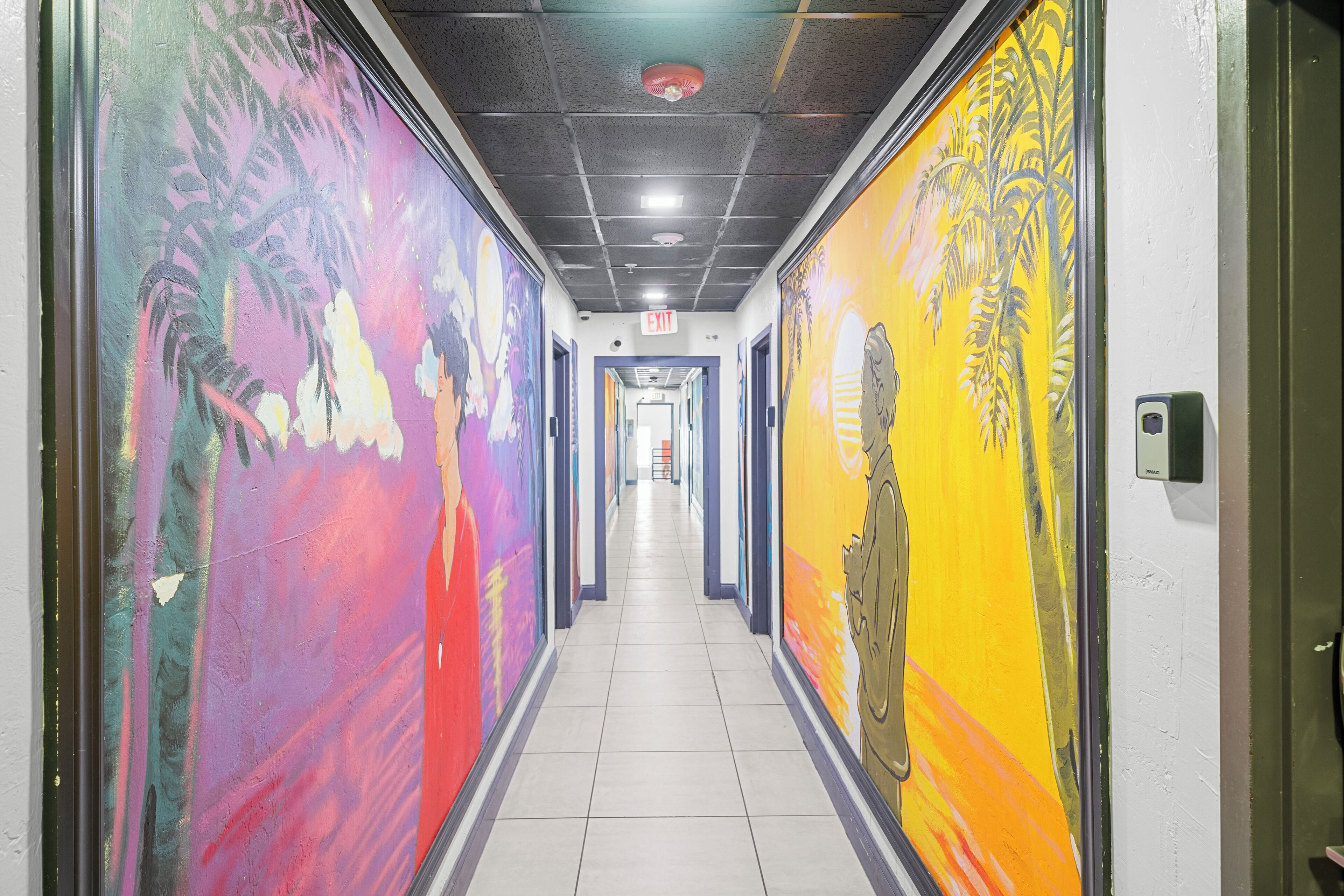 A hallway with colorful murals on the walls, a tiled floor, and an exit sign at the end.