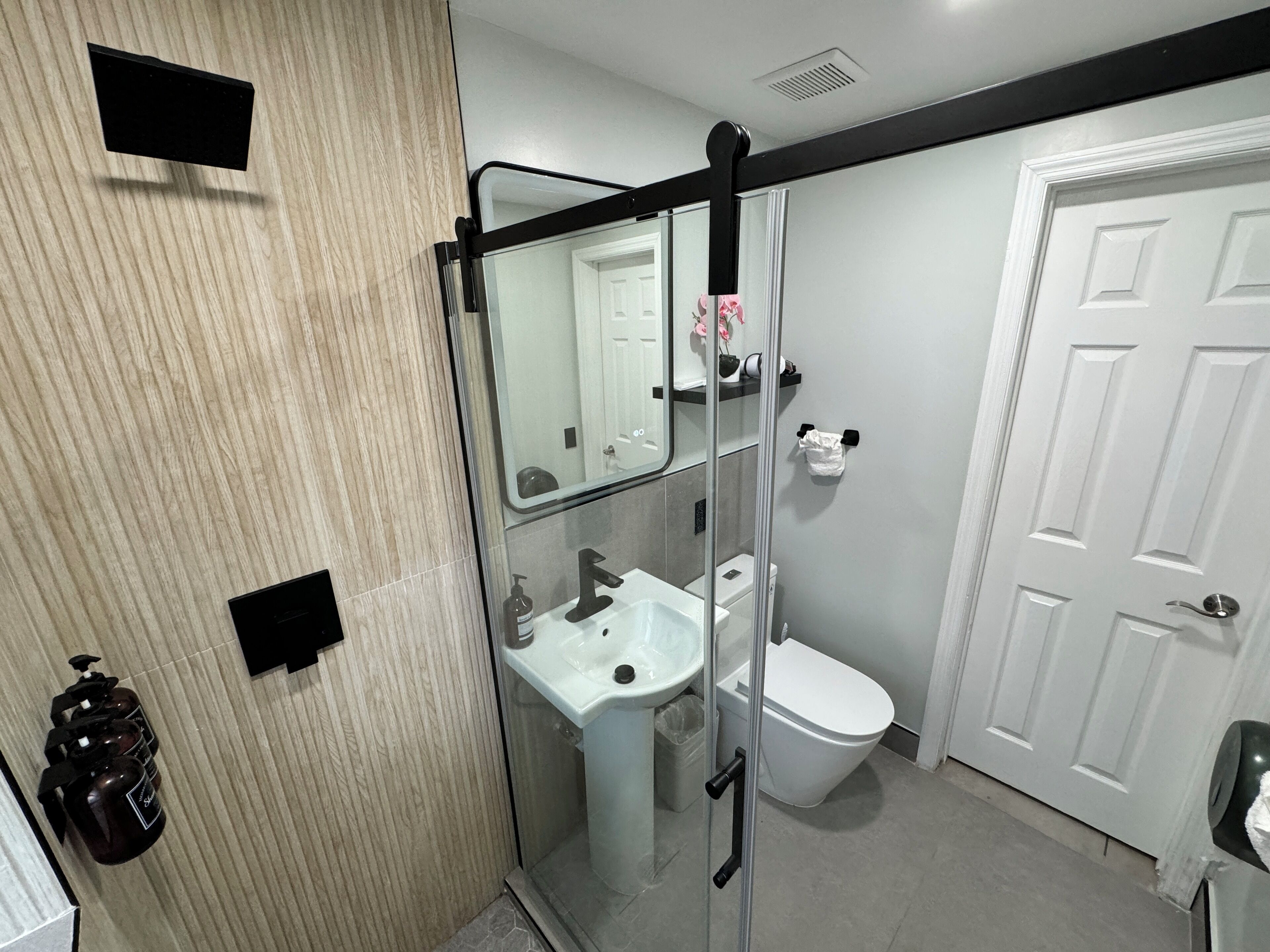 Une salle de bain avec une douche à parois de verre, un lavabo blanc, des toilettes et une porte.