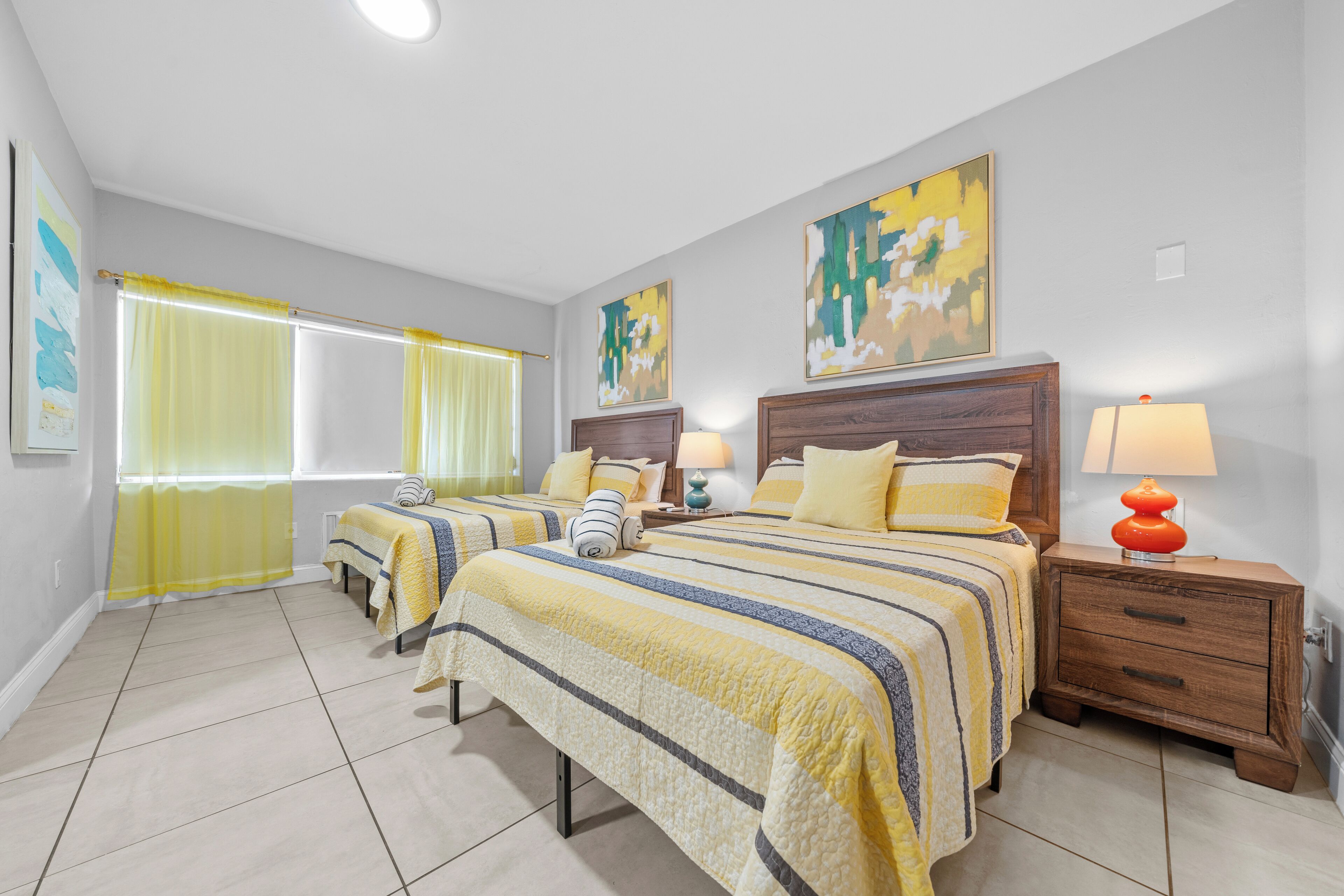 A hotel room with two beds, a nightstand with a lamp, and artwork on the walls.
