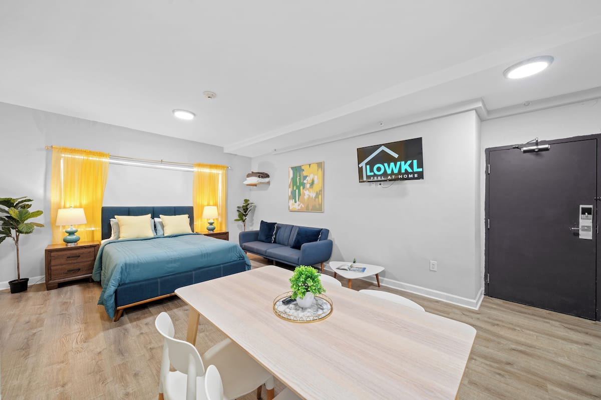 A modern hotel room with a bed, a sofa, a dining table, and a door.