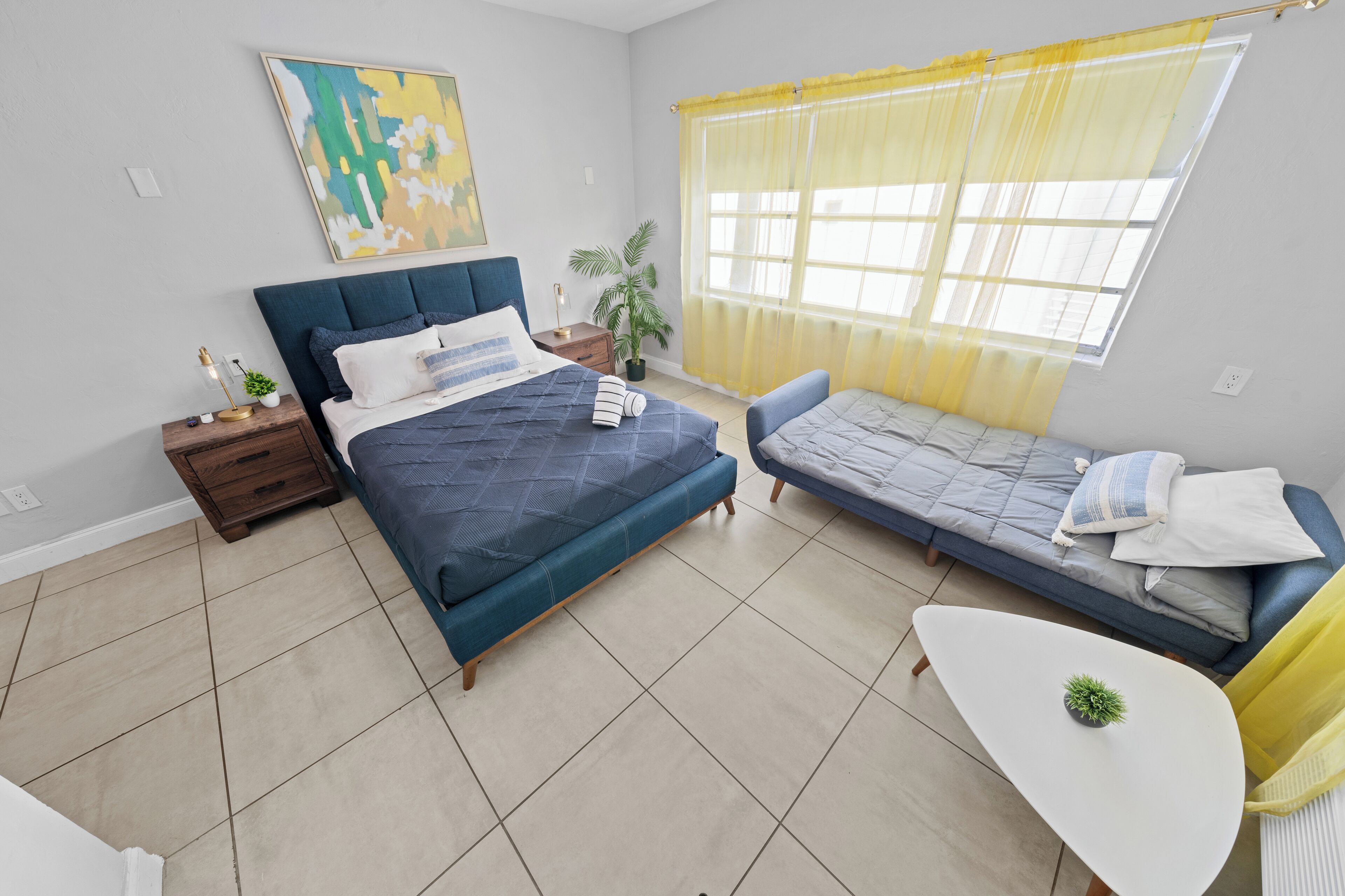 Une chambre à coucher comprenant un lit, un canapé, une petite table, une table de chevet, une plante et une fenêtre avec des rideaux.
