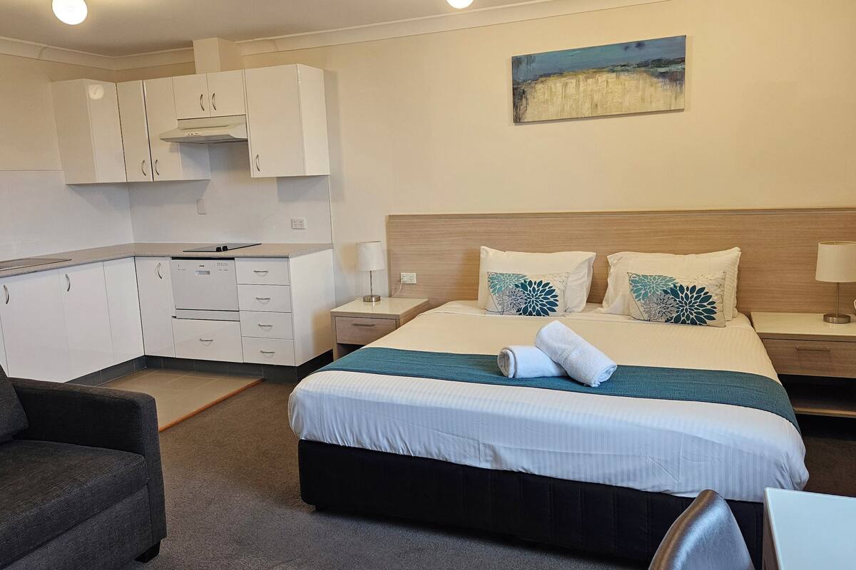 Quarto de hotel com cama, escrivaninha, cadeira, cozinha compacta e um quadro na parede.
