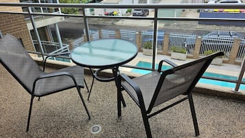 A balcony with a glass table and two chairs, overlooking a pool and a street with parked cars.
