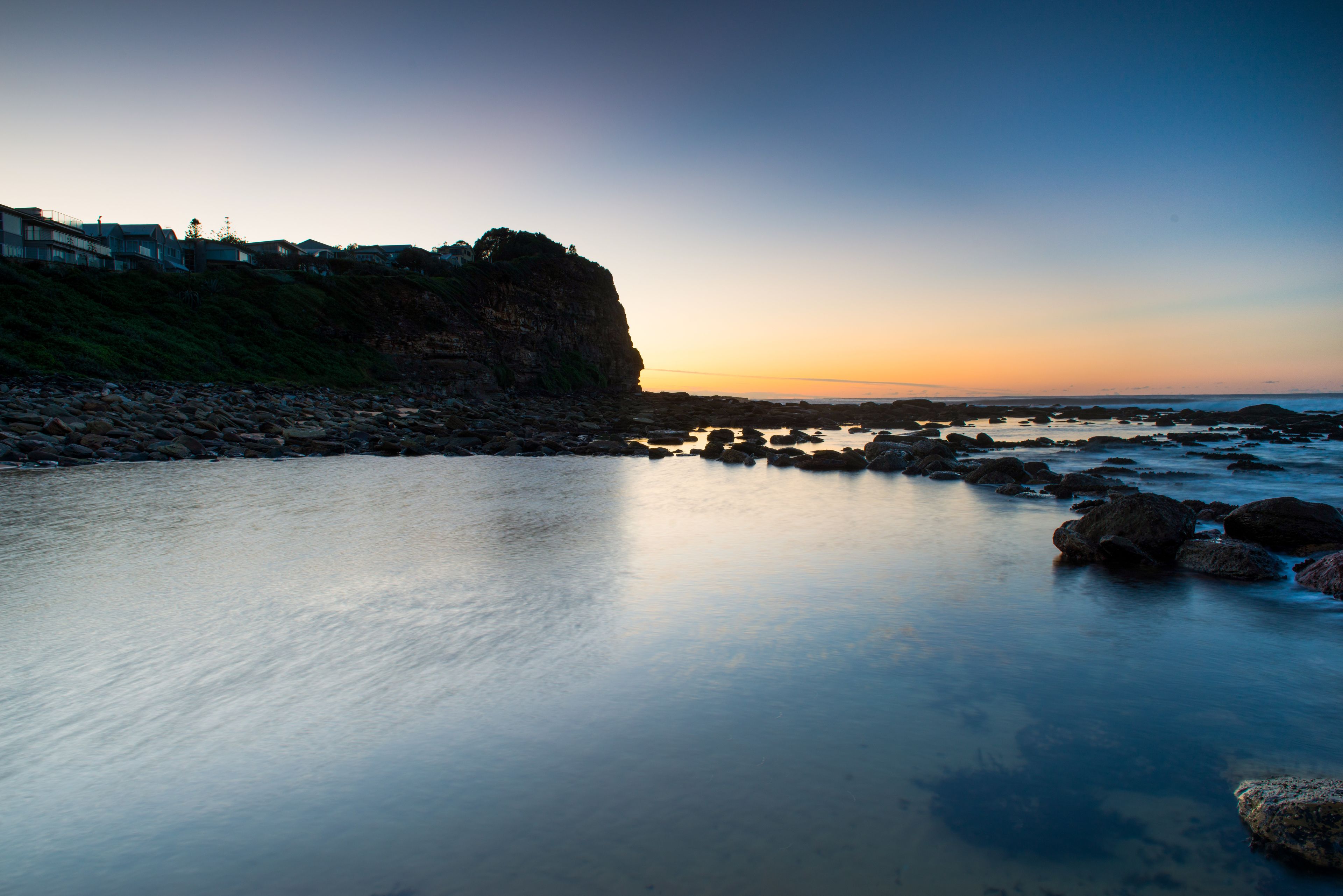 A coastal landscape with a rocky shoreline, calm water, and a cliff with buildings on top.