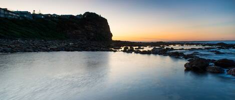 A coastal landscape with a rocky shoreline, calm water, and a cliff with buildings on top.