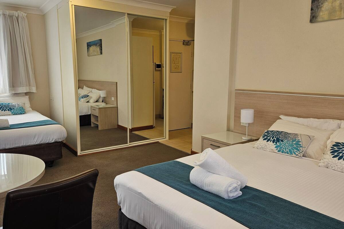 Quarto de hotel com duas camas, área de estar com mesa e cadeiras, um espelho e porta para o banheiro.