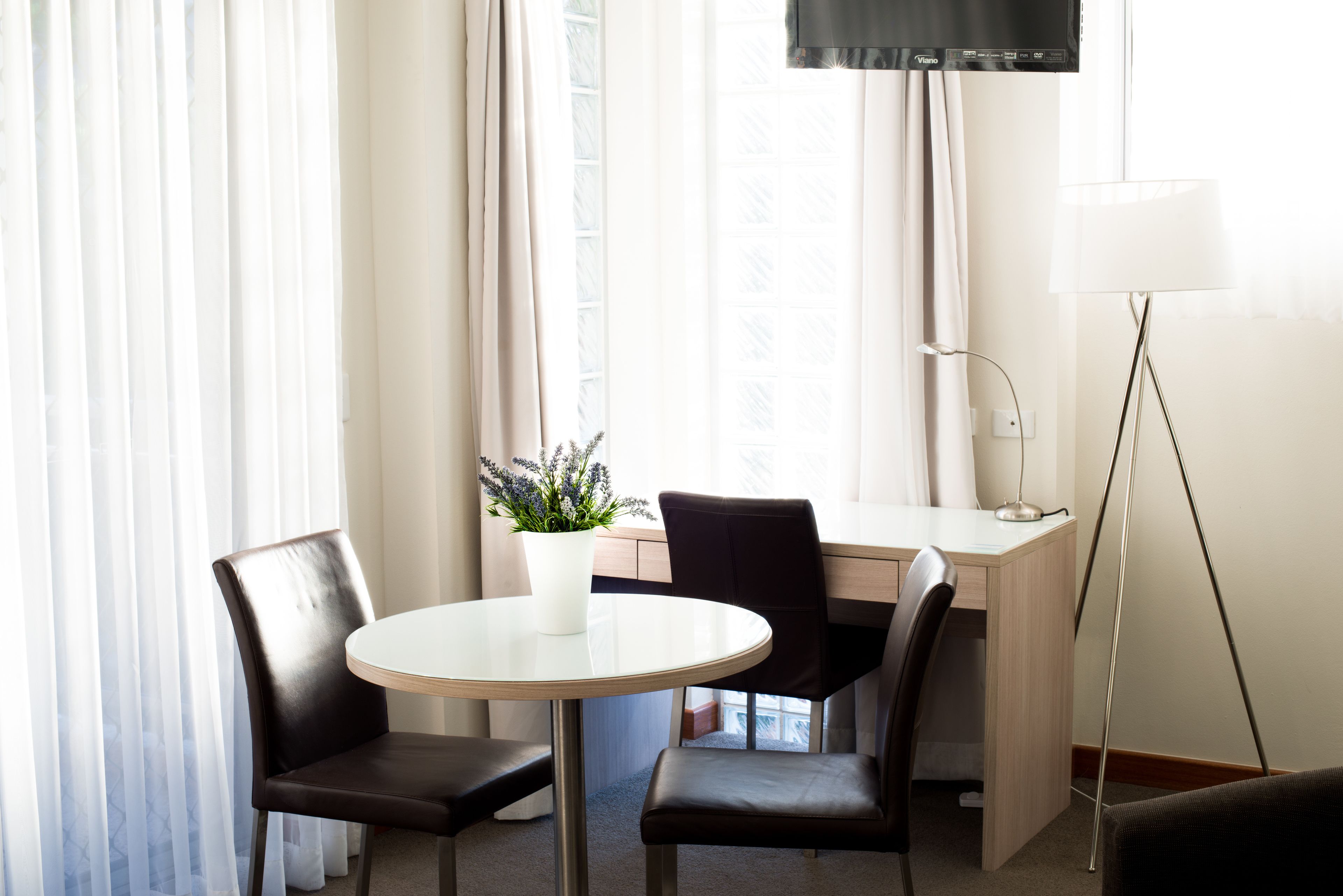 A hotel room with a round table, two chairs, a desk, a computer, a lamp, and a potted plant.