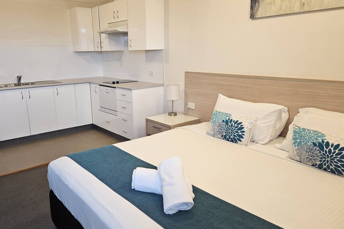 Quarto de hotel com cama, mesa de cabeceira, abajur, cadeira e cozinha compacta.