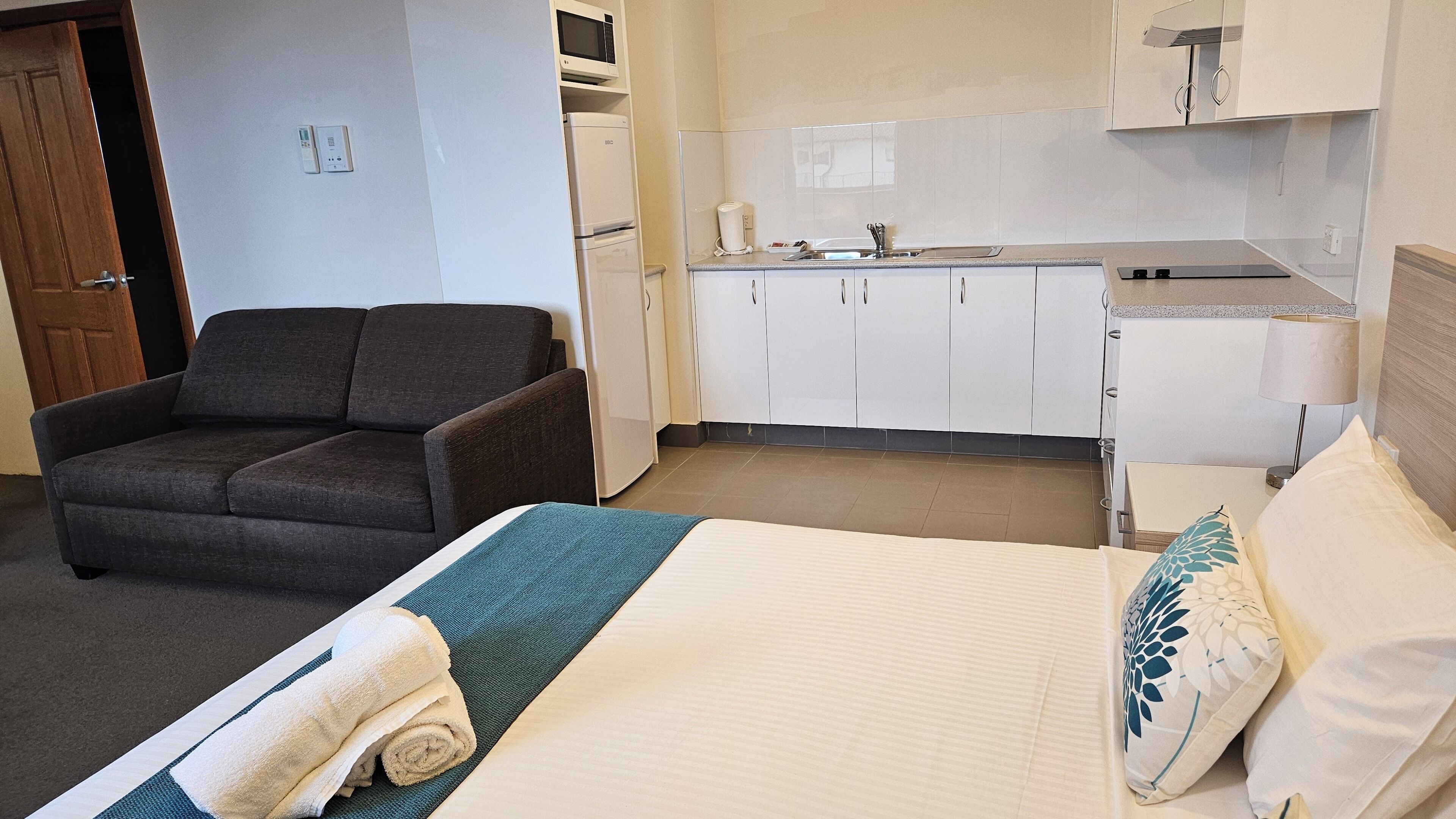 A hotel room with a bed, a chair, a kitchenette, and a door.