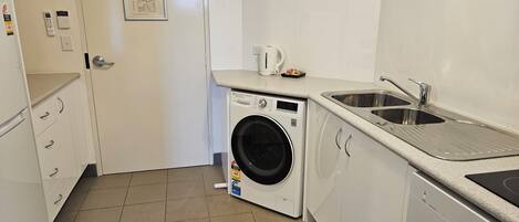 A compact laundry room with a front-loading washing machine, a sink, and a small countertop with a kettle.